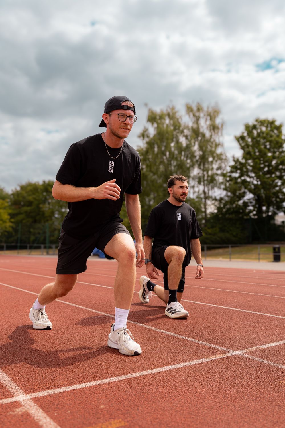 Two members doing walking lunges on a running track on a bright day